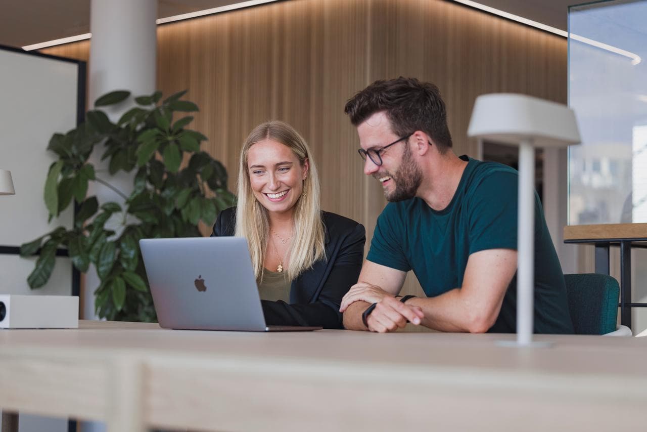 Two office workers, sitting at a table, smiling, both looking towards a laptop