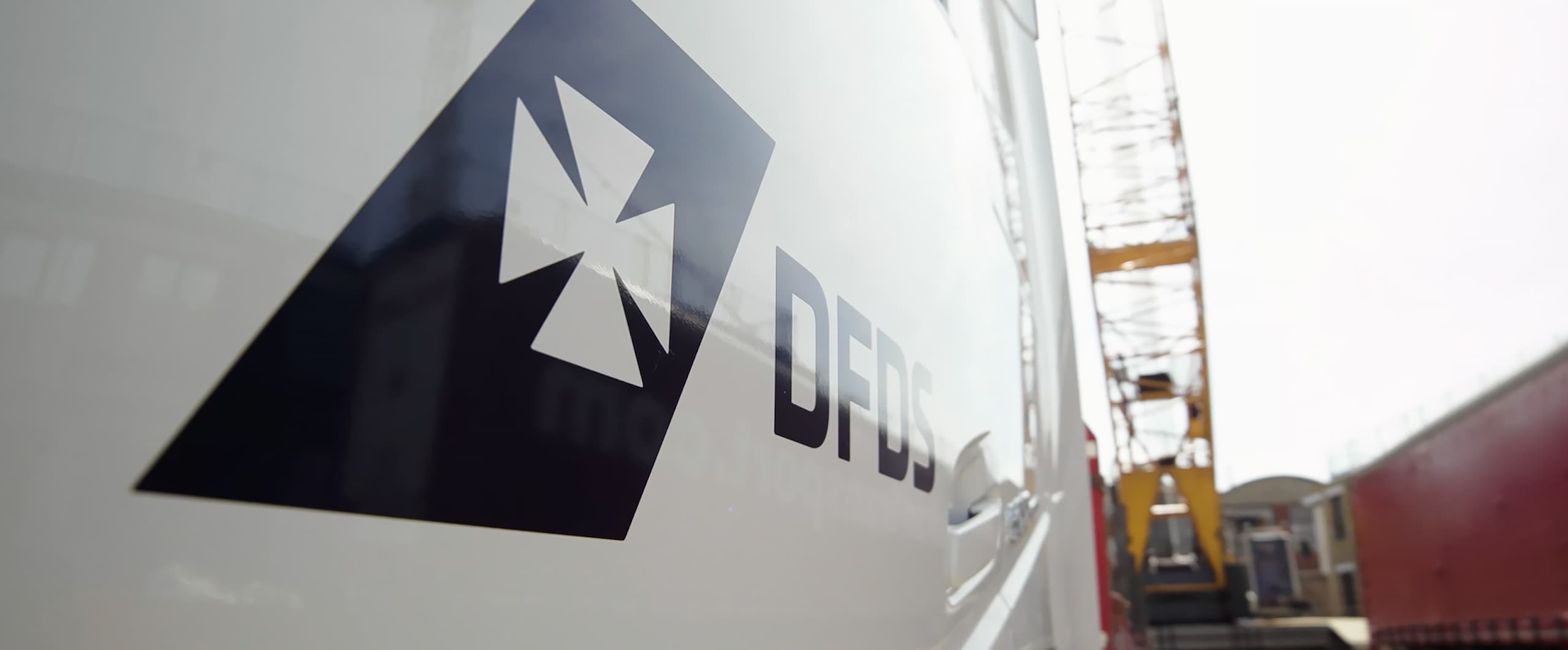 Close up of the DFDS logo on the side door of a truck, parked near other trucks and a crane