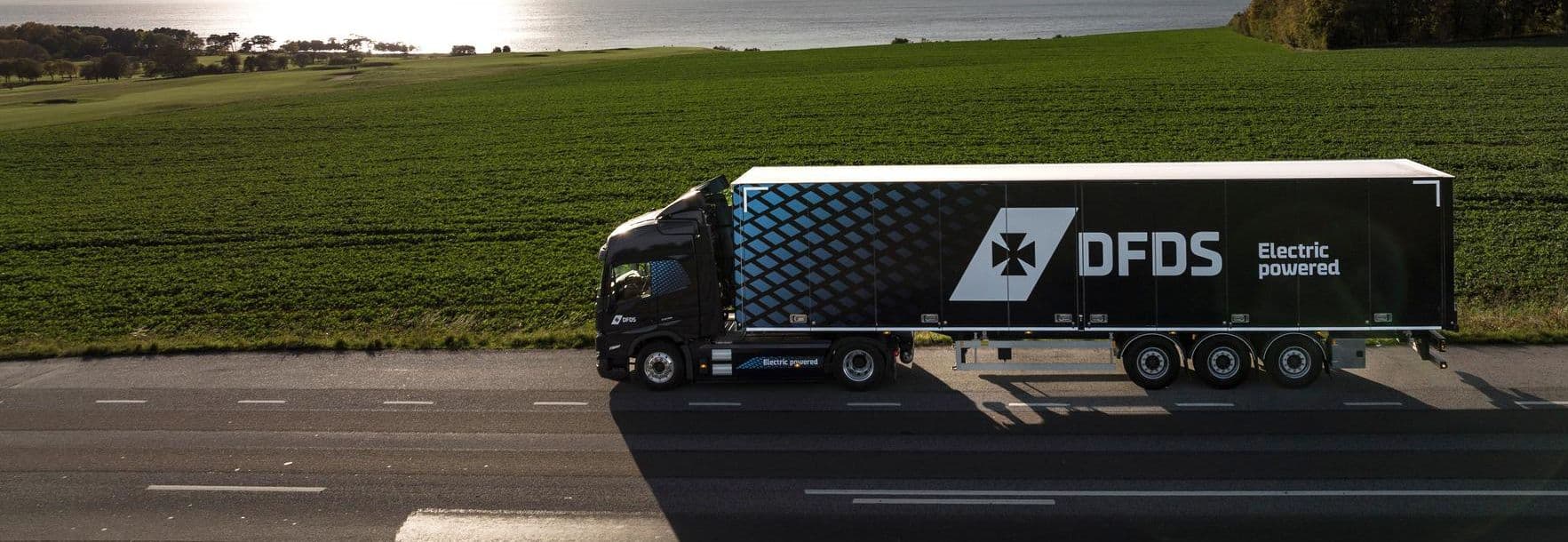 Blue electric truck with blue trailer driving on a road, with green surroundings