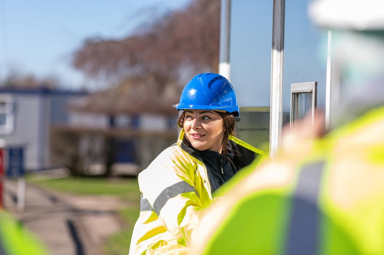 A woman standing outside in the sun, wearing a blue helmet and a yellow safety jacket, smiling and looking over her right shoulder backwards
