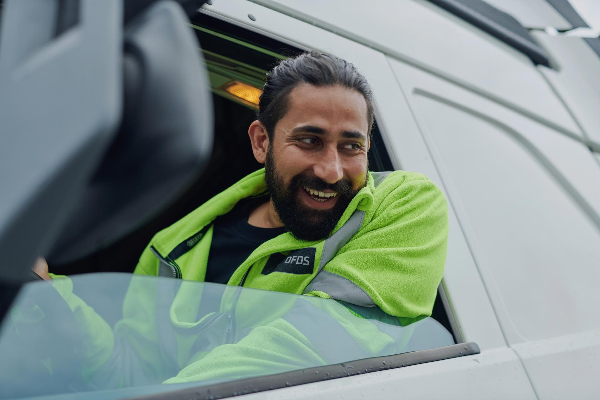 A male driver in white DFDS truck, wearing yellow safety jacket, leaning out of window, smiling