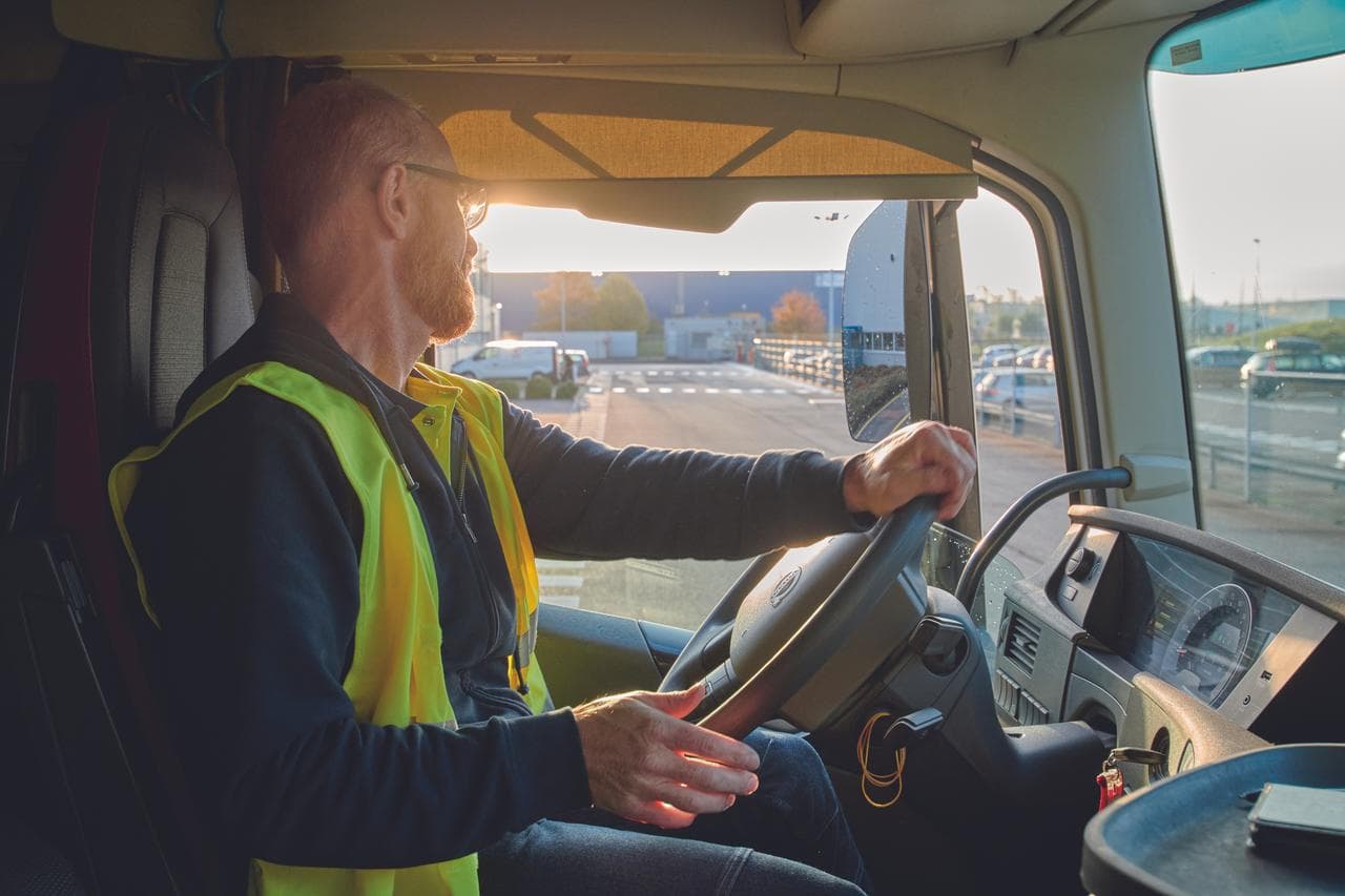 An inside picture of a truck with a driver sitting behind the steering, wearing a yellow security vest