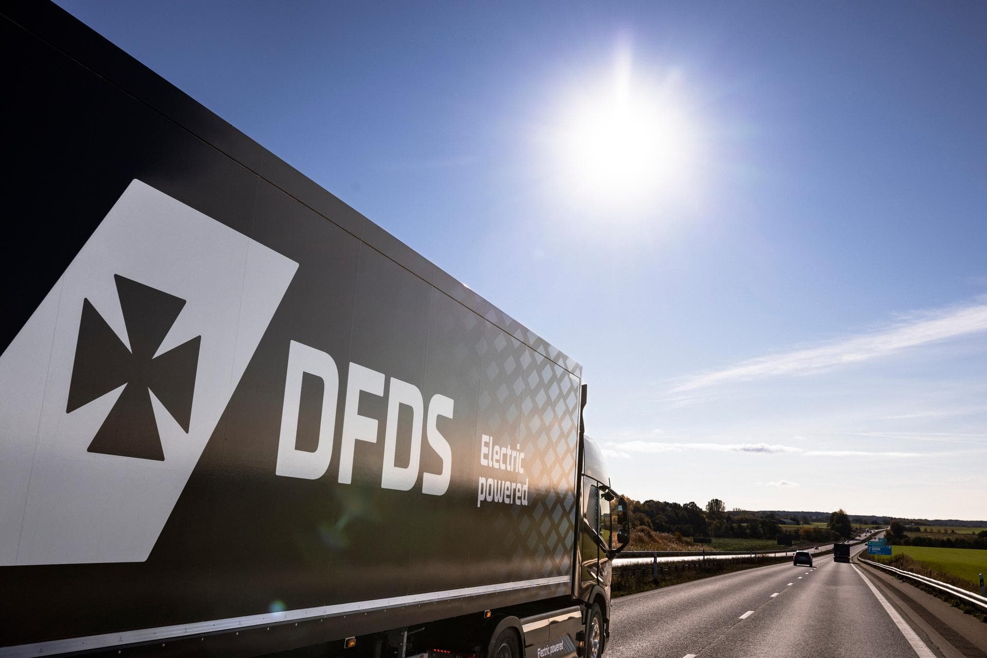 A dark blue DFDS trailer with an electric truck seen from the side, driving on the quiet highway, with the bright sun up in the sky
